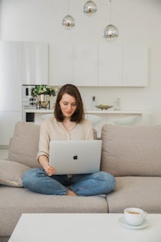 Young woman comfortably using a laptop on her sofa in a bright living room.