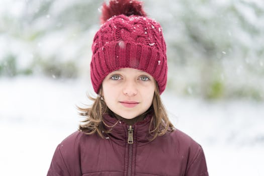 Calm young girl with short brown hair and light eyes in winter down jacket and hat with pompom standing on street with falling snowflakes in winter park