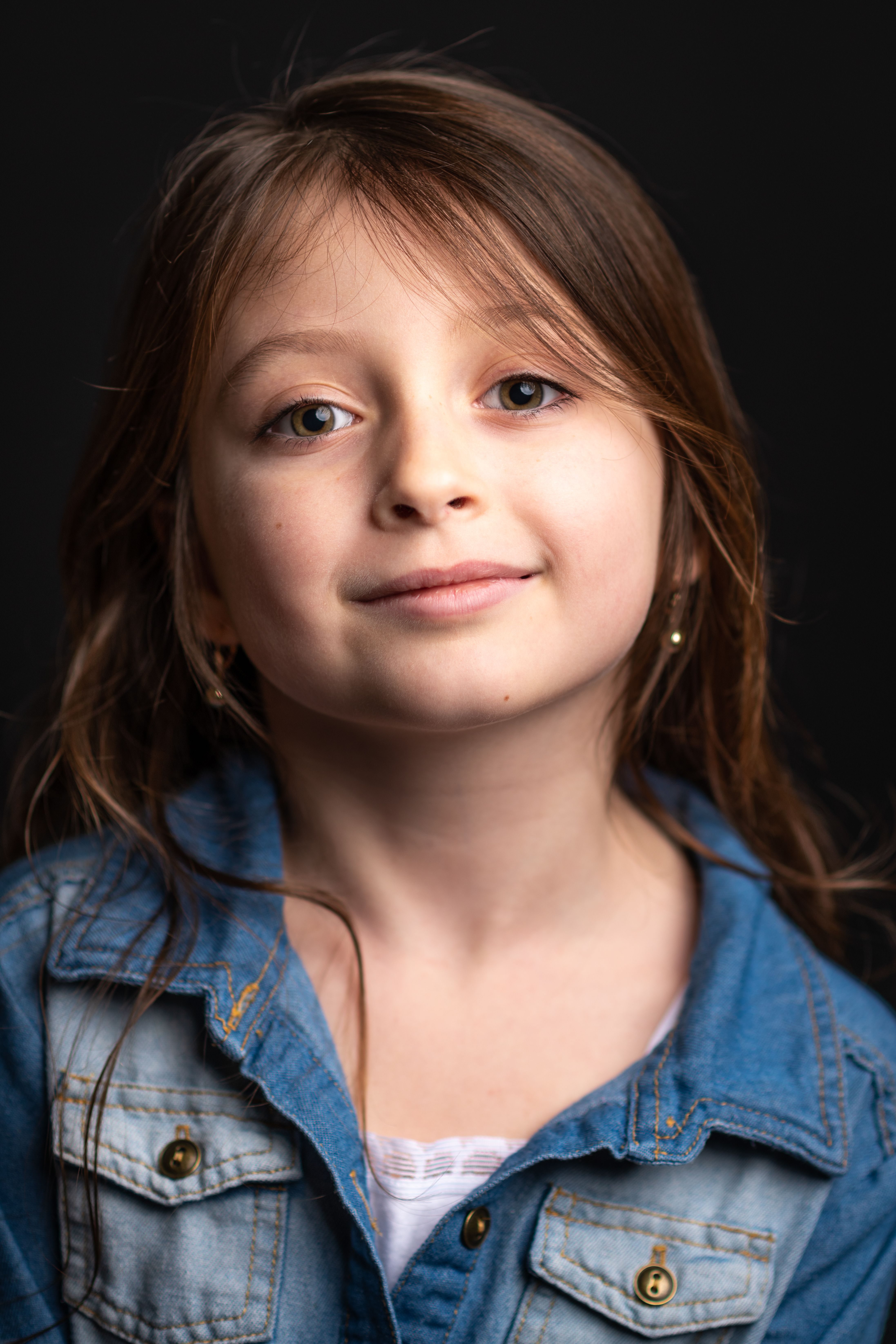Charming young girl with long hair and blue denim jacket smiling confidently in a studio portrait.