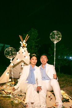 晚上，一对穿着白色西装的时尚亚洲夫妇坐在明亮的帐篷外