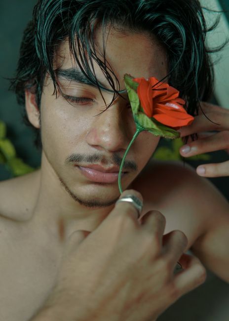 Thoughtful young ethnic man with eyes closed covering face with red flower while sitting with bare chest