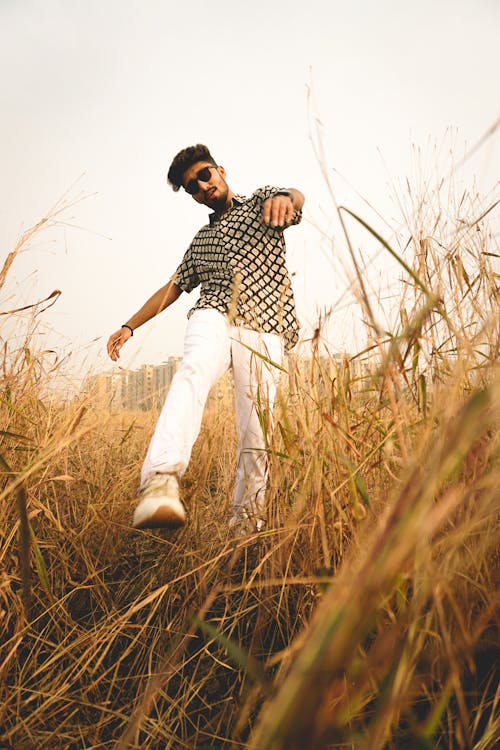 Low angle full body of calm young male in stylish outfit and glasses walking over grass while enjoying nature