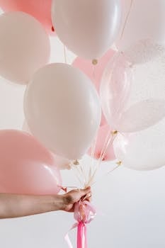A pastel pink and white balloon bouquet held by a hand against a light background, ideal for celebrations.