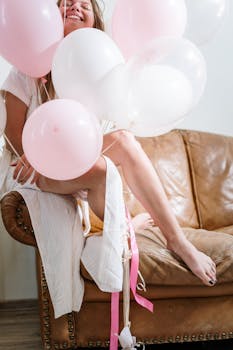 A young woman celebrates joyfully with pastel pink and white balloons indoors on a leather sofa.