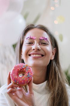 一位快乐的女子拿着一个五颜六色的甜甜圈，展示着节日妆容和灿烂的笑容