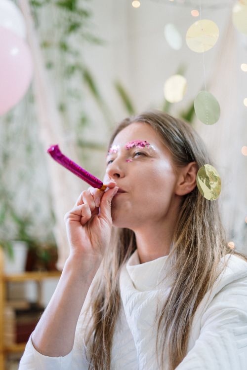 Free A woman with glitter makeup indoors blowing a pink party horn at a celebration. Stock Photo