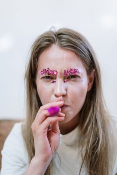 A whimsical portrait of a woman with glitter makeup, playfully blowing a party horn indoors.