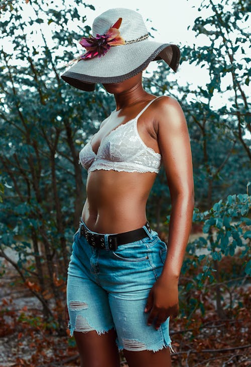 Sensual young African American faceless female showing body while covering face with large hat