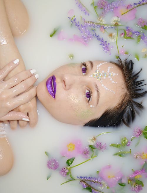 Free From above of relaxed female with rhinestones on forehead looking up while lying in bathtub with purple flowers and twigs during skincare routine Stock Photo