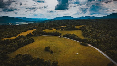 Drone view of wavy roadway between grass meadows with woods behind mounts under cloudy sky