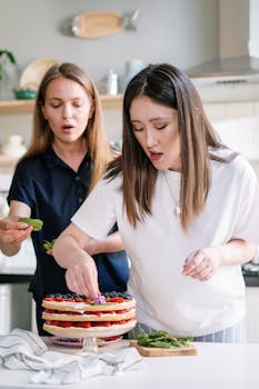 Two women are decorating a fresh cake with berries in a bright, modern kitchen setting.