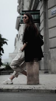 Young female with dark hair in stylish clothes and sunglasses holding hands in pockets and sitting on concrete construction on street at daytime