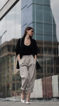 Low angle full body of confident female with hands in pockets wearing trendy clothes walking near contemporary glass facades