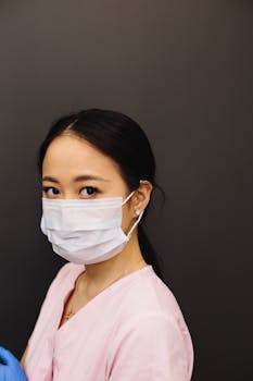A focused portrait of a healthcare worker in a pink uniform wearing a face mask, standing indoors.