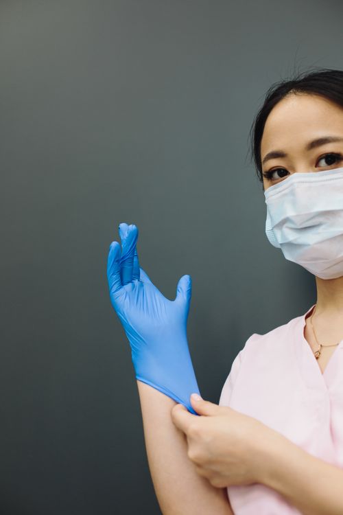 Free Asian woman in medical mask putting on gloves, symbolizing hygiene and safety. Stock Photo