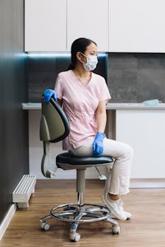 Healthcare worker in a clinic wearing a face mask and gloves, seated on a chair, symbolizing modern healthcare.