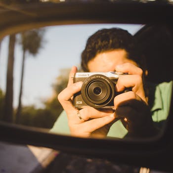 A close-up of a man taking a photo through a car's side view mirror.