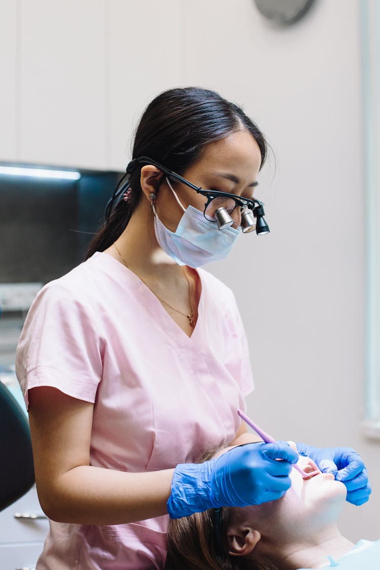 Dentist In Pink Scrub And Black Framed Eyeglasses With Loupe Examining A Patient