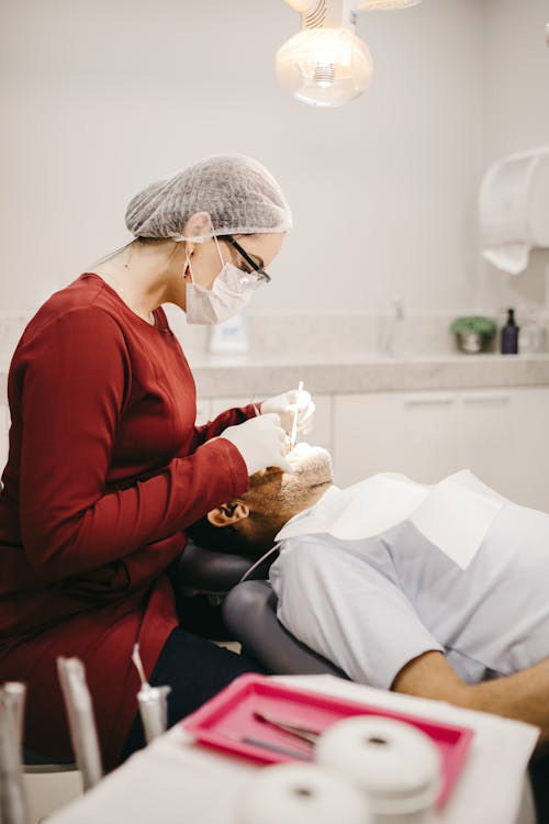 Side view of anonymous female stomatologist in eyeglasses and gloves checking teeth of lying man in clinic