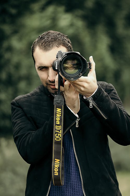 Concentrated male photographer in casual clothes looking through objective lens of camera while taking photo on nature with blurred background