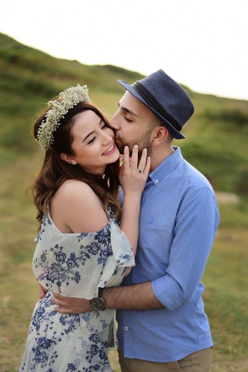 Tender man in hat kissing charming woman with wreath on cheek while hugging on grassy meadow in daylight in nature