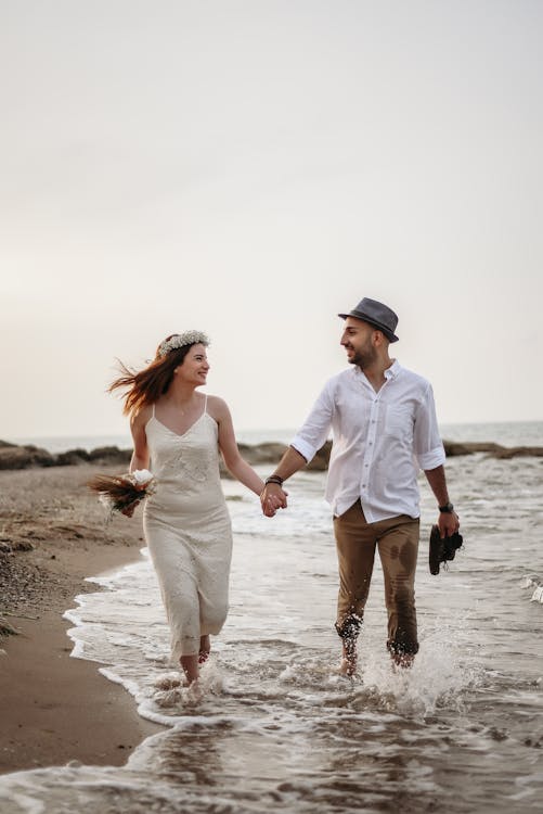 Free Loving couple in white clothes and headdresses running on coastline in summertime against gray sky and looking at each other while holding hands Stock Photo