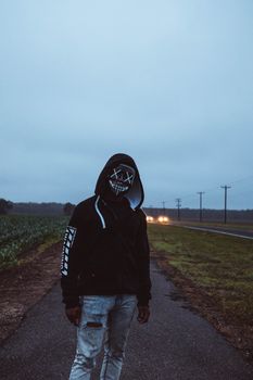 黄昏时分，一个戴着发光面具和连帽衫的人站在一条薄雾弥漫的乡村道路上