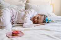一位开朗的成年人穿着睡衣，戴着甜甜圈和派对帽在床上庆祝生日