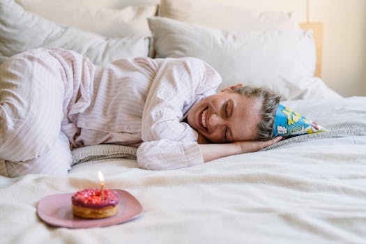 A cheerful adult in pajamas celebrates a birthday morning in bed with a donut and party hat.