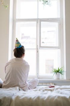 A woman in pajamas with a birthday hat sits alone, indoors by a window.