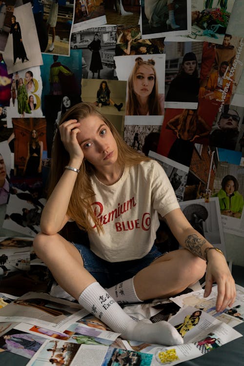 Young girl with tattoo on arm in casual clothes siting on homemade wallpaper made from cut out pages of magazine