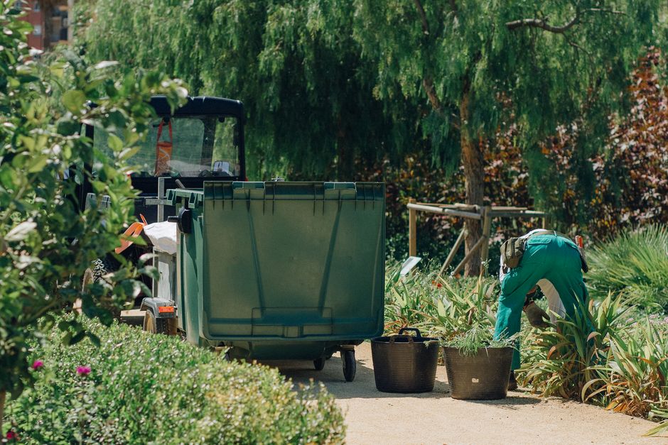 A gardener tends to plants beside a large green bin in a lush park