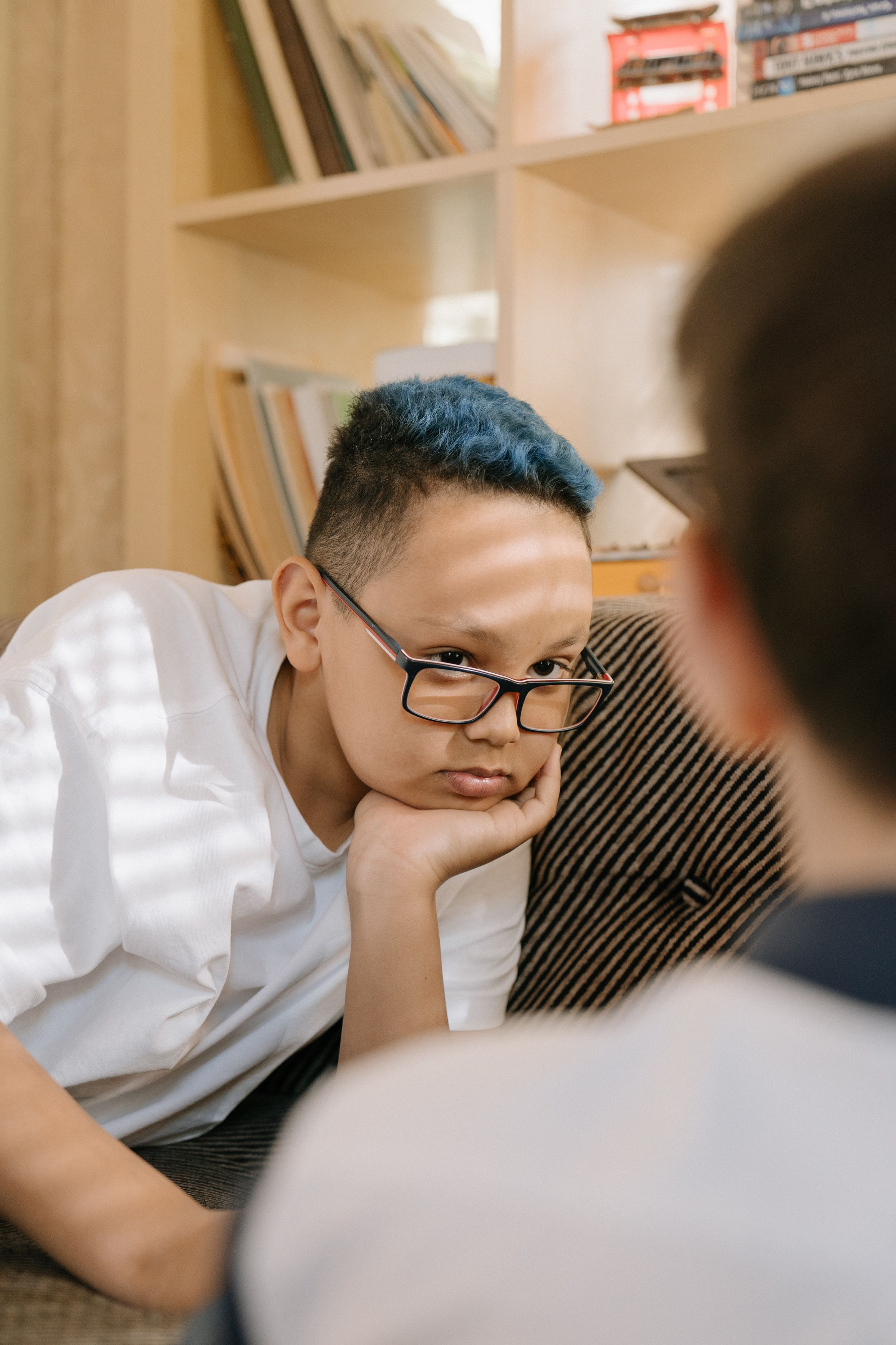 一个蓝头发、戴眼镜的小男孩，在室内用手托着下巴沉思