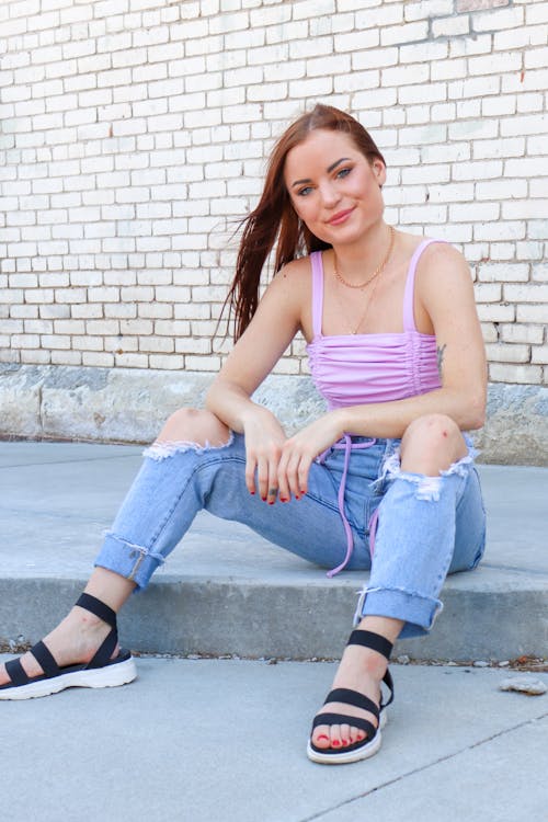 Free Woman in Purple Tank Top and Ripped Denim Jeans Sitting on Sidewalk Stock Photo