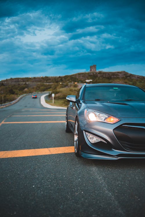 Vibrant blue sky covered with clouds over dark grey luxury sports car riding on asphalt road with turned headlights