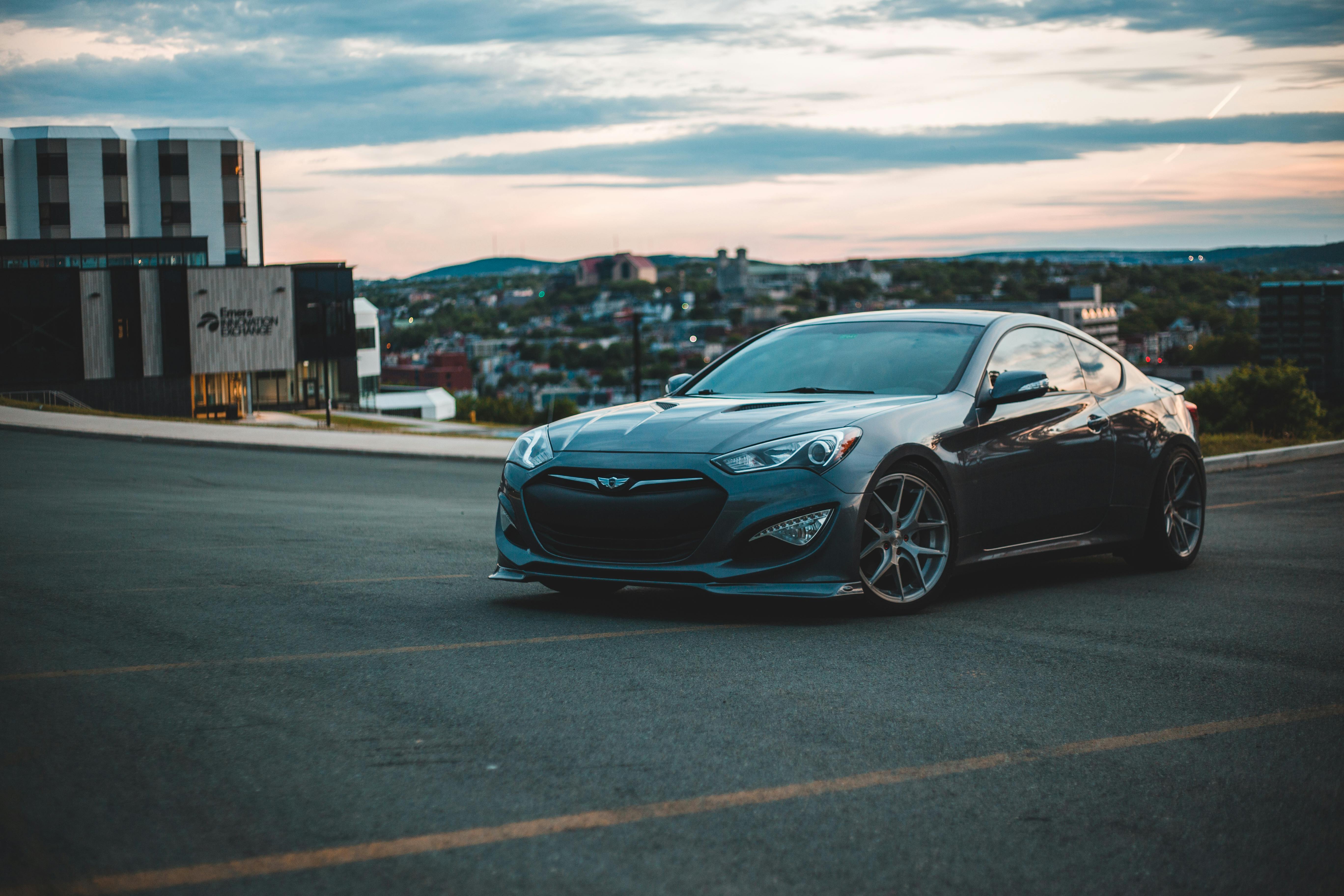 Black expensive sports car with glossy surface parked on paved car parking next to building and settlement under light blue evening sky covered with clouds
