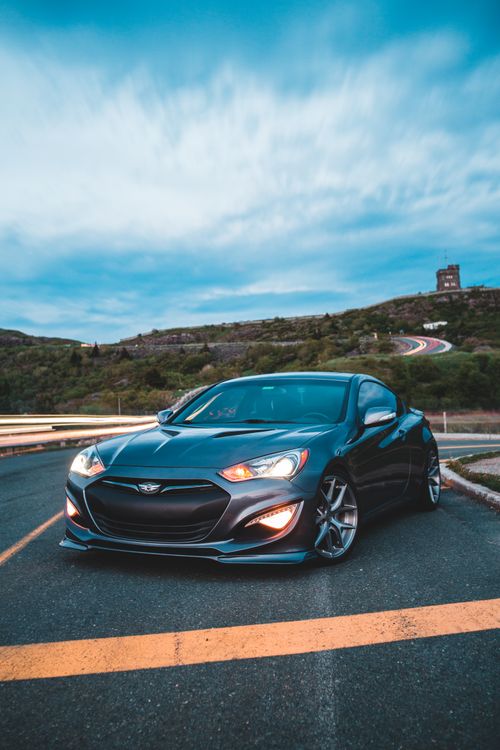 Free Luxury black vehicle with glossy surface and turned red headlights on paved road near slopes covered with grass and tiny tower on horizon in Canada Stock Photo