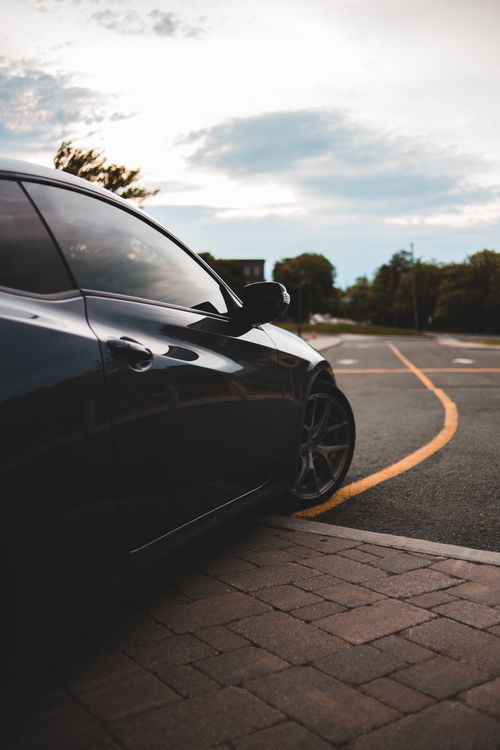 Free Side part of black luxury automobile with glossy surface parked on pavement near asphalt road under light pink evening sky Stock Photo