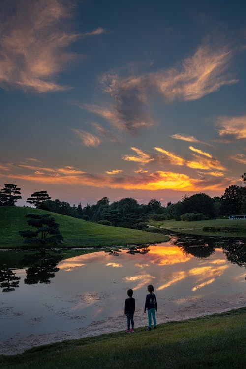 High angle full body back view of anonymous children enjoying picturesque view of calm river flowing through hilly terrain in evening