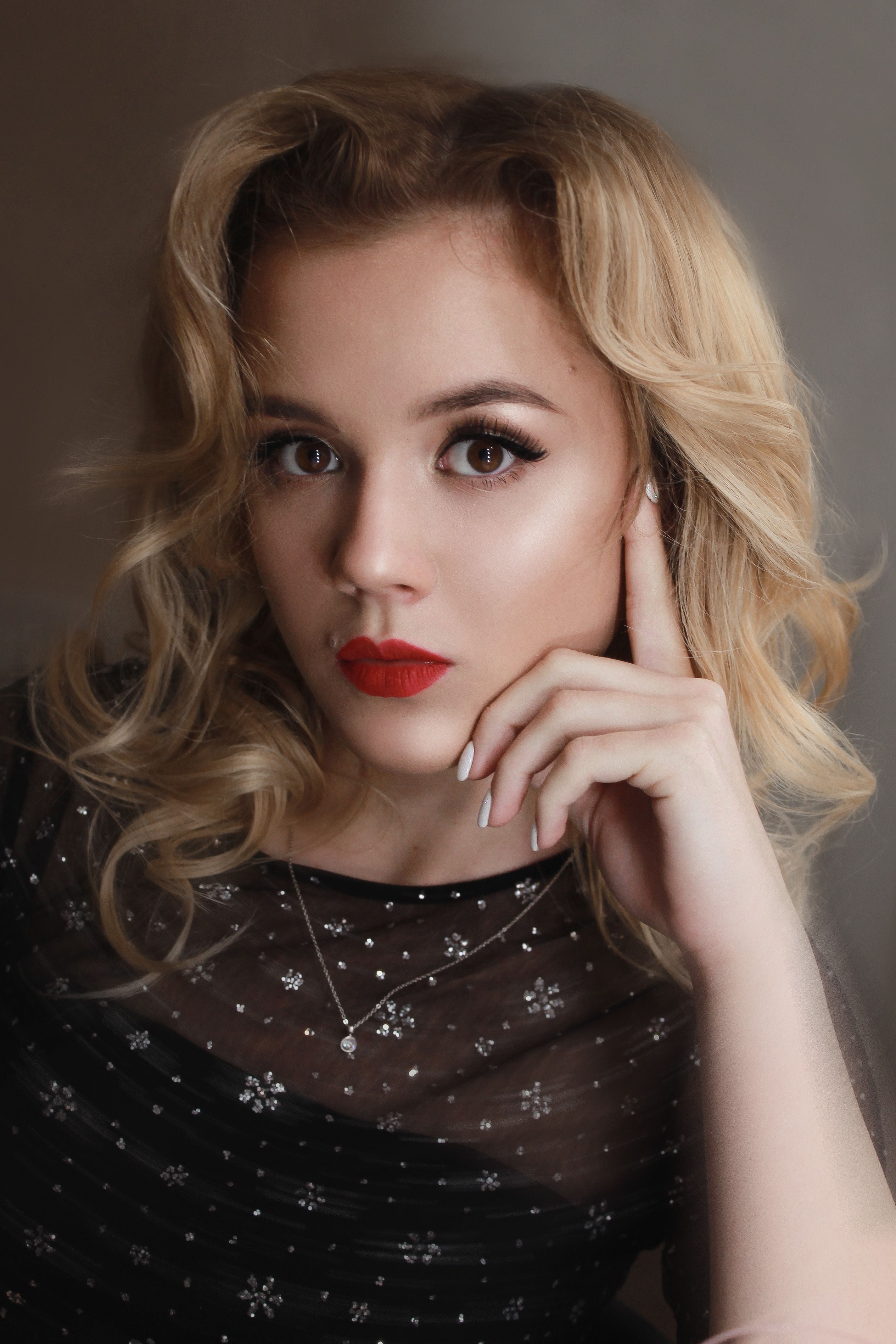 Charming woman in black outfit with blond hair and red lipstick looking at camera while leaning on hand against gray background