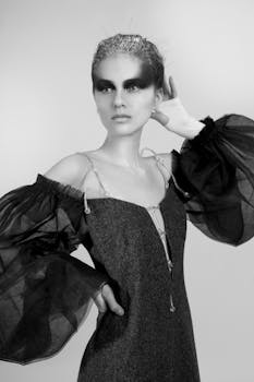 Black and white of sensual female with bright makeup wearing dress with naked shoulders and transparent sleeves standing with hand on waist