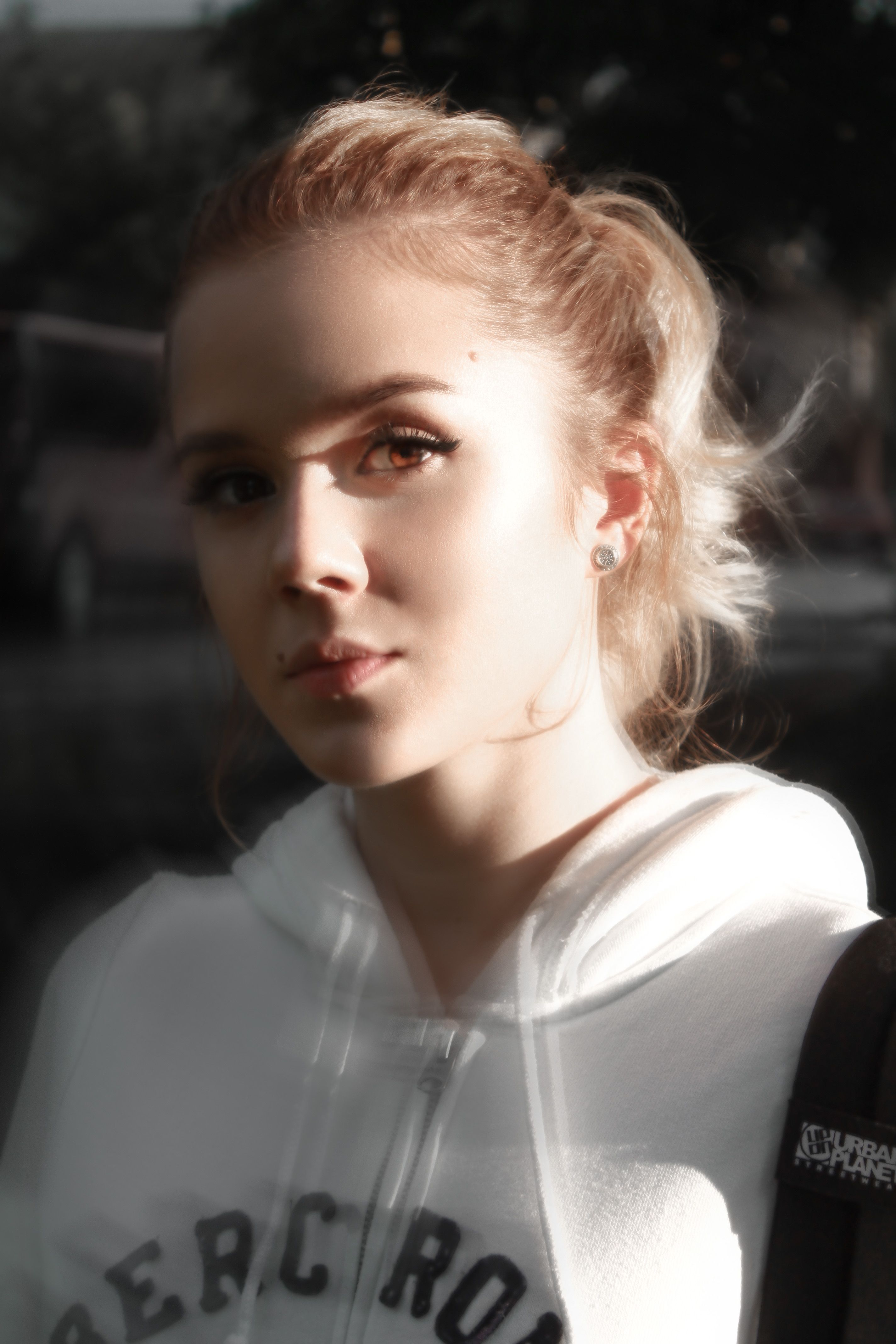 Pleased female with blond hair in ponytail wearing white sweatshirt standing on city street in sunlight while looking at camera