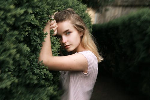 Content lady standing with hands on green shrub and looking at camera while standing in garden on blurred background in daylight