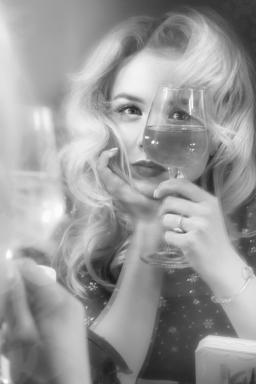 Free Black and white of cheerful woman with curly hair sitting with chin on hand and looking through wineglass in hand Stock Photo