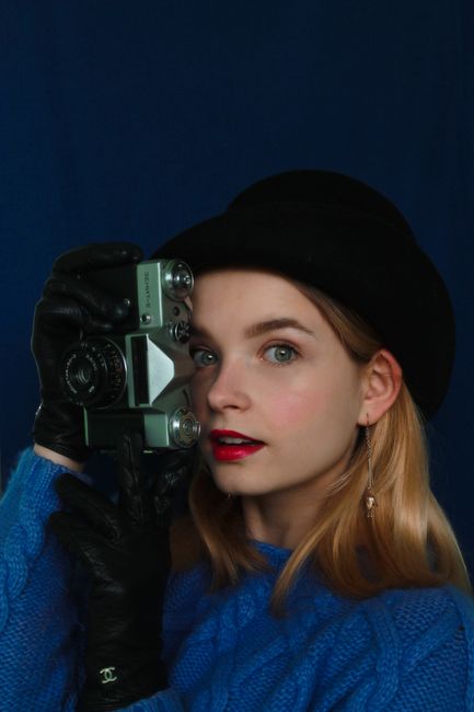 Content lady in blue outfit wearing hat and gloves looking through lenses of retro camera while standing on blue background in studio