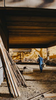 A worker in blue uniform at a construction site under an overpass, carrying equipment.
