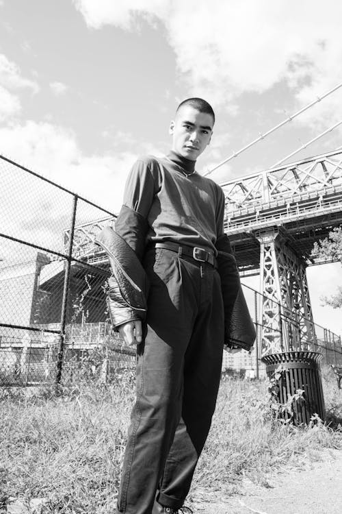 Grayscale Photo of Man in Long Sleeve Shirt and Pants Standing Near Metal Fence