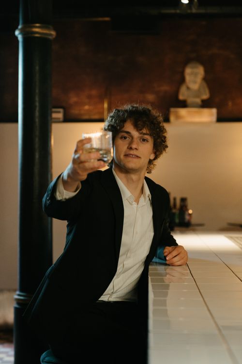 Free A young man in a suit raises a drink at a stylish bar counter with a statue backdrop. Stock Photo
