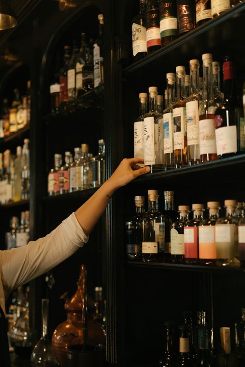 Free A hand reaching for bottles on a bar shelf, showcasing a variety of spirits in an elegant indoor setting. Stock Photo