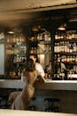 一名女子优雅地坐在摆满瓶子的现代酒吧柜台前，与调酒师聊天
